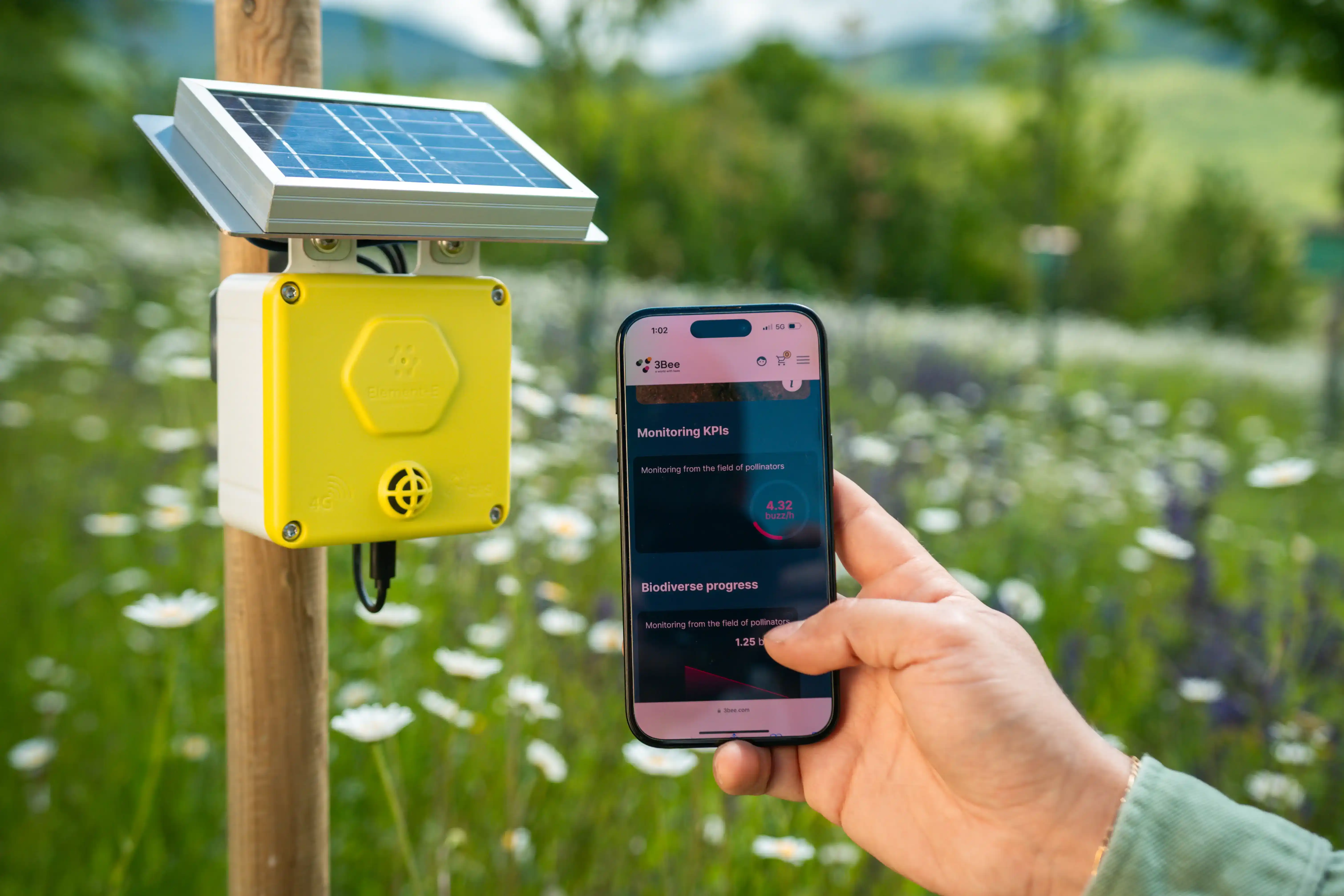 A close-up of a hand holding a smartphone in front of a solar-powered Spectrum device mounted on a wooden post in a flower field. The smartphone screen shows an app displaying monitoring KPIs and biodiversity progress